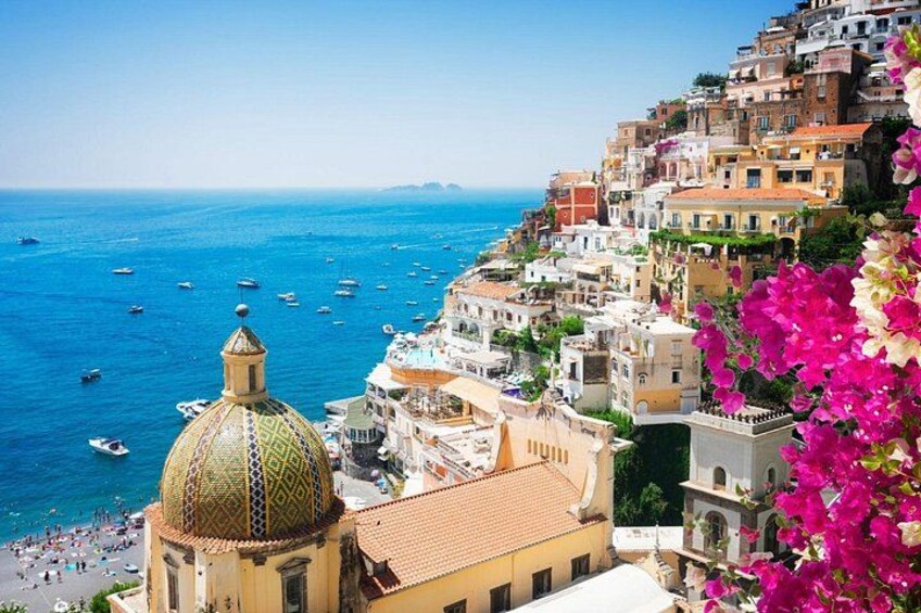 A coastal town with a prominent golden-domed building, a densely packed hillside with colorful buildings, and a clear blue sea with boats. 1