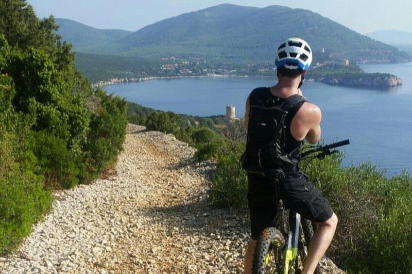 Una persona en bicicleta de montaña, con casco, pedaleando por un camino de grava con un lago y montañas al fondo. 1