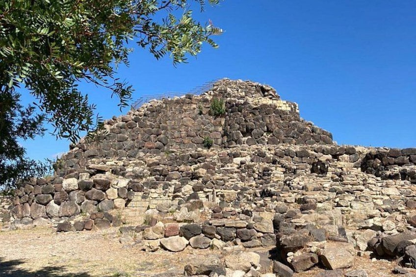 A large, ancient stone structure with a flat top and steep sides, surrounded by dry grass and a clear blue sky. 3