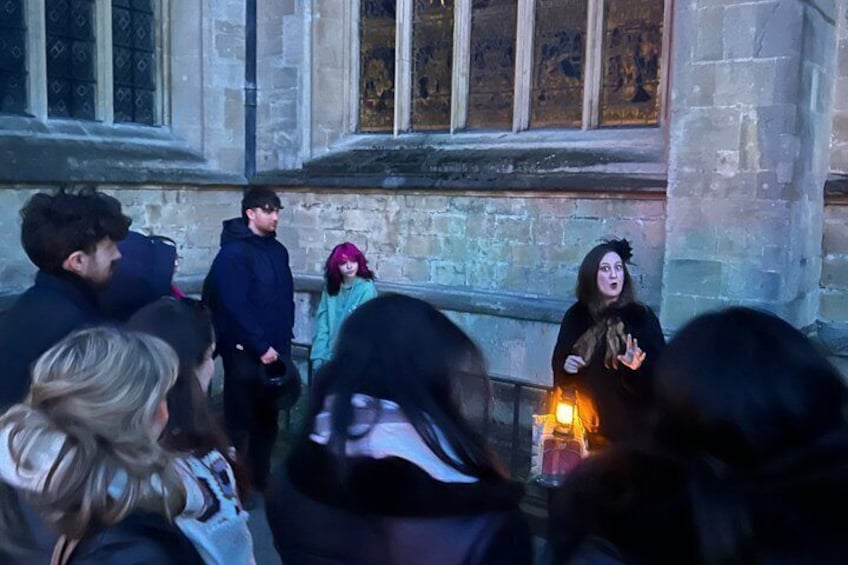 A group of people gathered around a person holding a candle in front of a stone building with large windows. 4