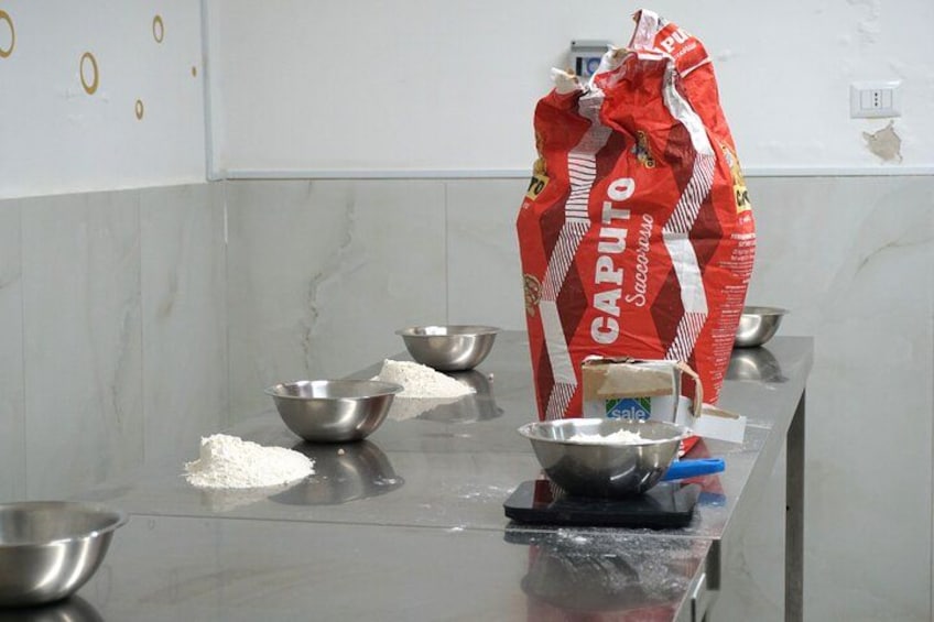 A kitchen counter with a bag of Caputo flour, several metal bowls, and a blue container. 3