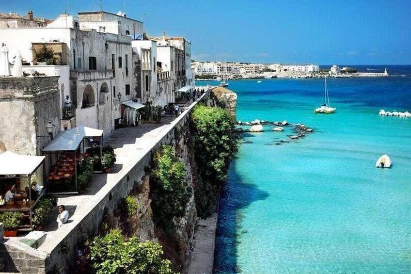 A coastal town with buildings along a cliff overlooking turquoise waters, boats, and a clear sky. 3