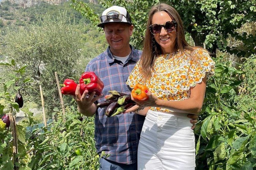 Two people holding vegetables in a garden with various plants and trees in the background. 4