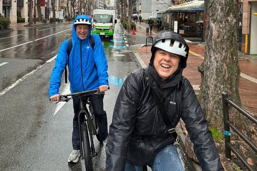 Dos personas en bicicleta, una con chaqueta azul y la otra con chaqueta negra, en una calle mojada de la ciudad. 4