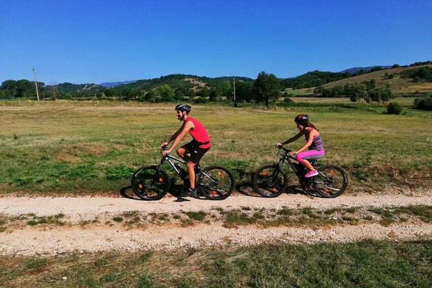 Two cyclists on a dirt path with grassy fields and hills in the background. 3
