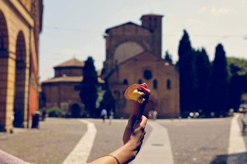 A hand holding a cone with a red topping, a historic building with a bell tower, and a clear sky. 2