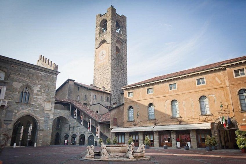 Una plaza histórica con una torre destacada, edificios de piedra y una fuente. 2