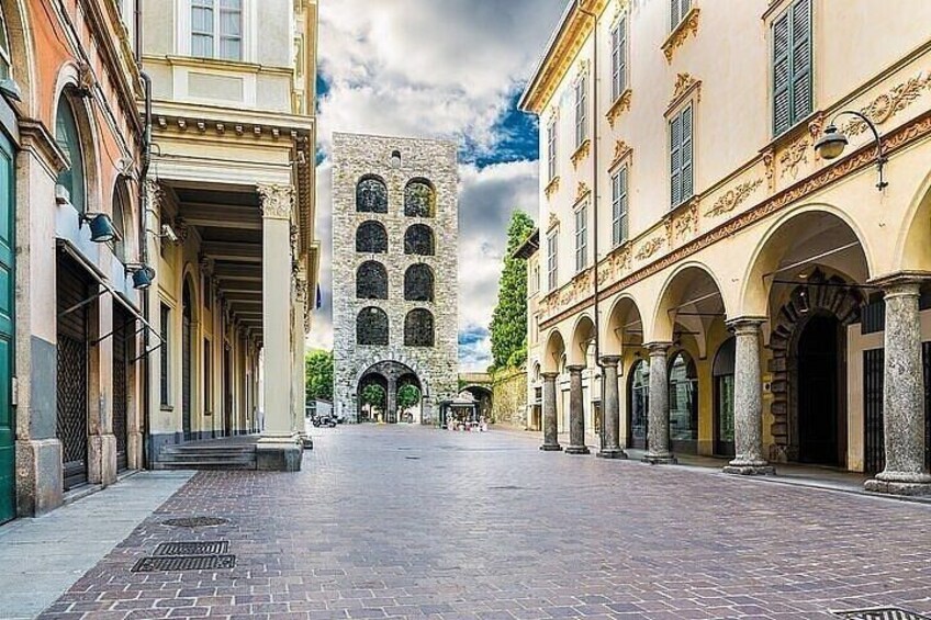 A cobblestone street with historic buildings, a central tower, and an open square. 5