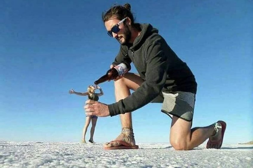 Una persona arrodillada sobre una superficie blanca y plana, sosteniendo una botella, con otra persona de pie al fondo. 2