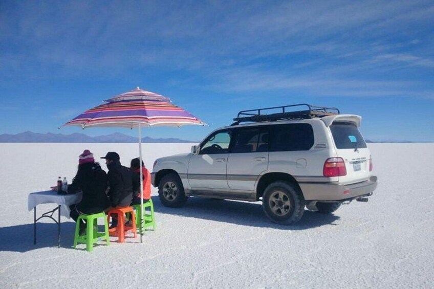 Een witte SUV geparkeerd op een zoutvlakte, met een kleurrijke strepen paraplu en mensen zittend aan een tafel. 2