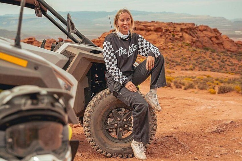 A person sitting on a vehicle's tire in a desert landscape with mountains in the background. 5