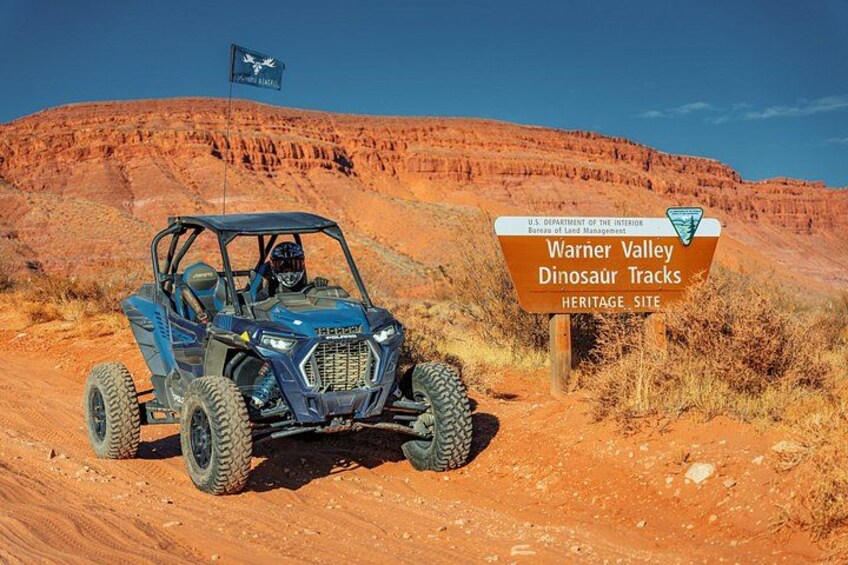 Un véhicule tout-terrain bleu sur un chemin de terre, avec un panneau indiquant le site patrimonial des empreintes de dinosaures de Warner Valley. 1