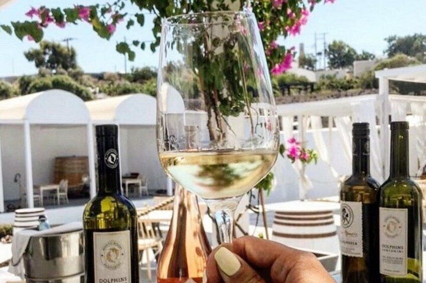 A glass of white wine held by a hand, with bottles and a white outdoor setting in the background. 2