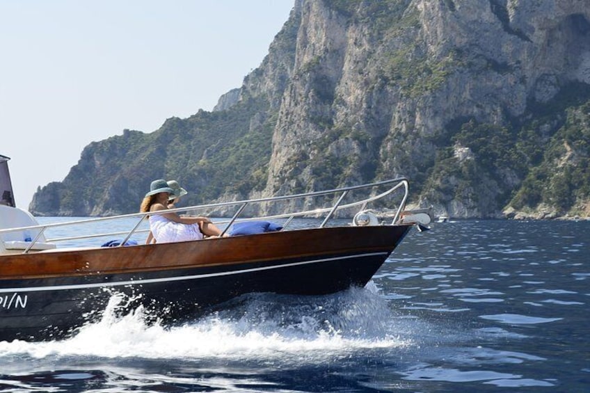 A wooden boat with two people on board, sailing on a calm sea with a rocky coastline in the background. 4