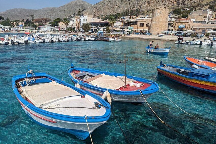A marina with several small boats docked in clear blue water, a castle in the background, and a hilly landscape. 3