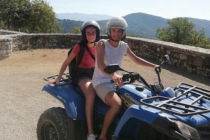 Two people on a blue ATV with a stone wall and mountains in the background. 1