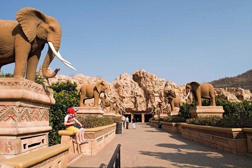 A pathway lined with elephant statues, a person sitting on a bench, and a rocky background. 3