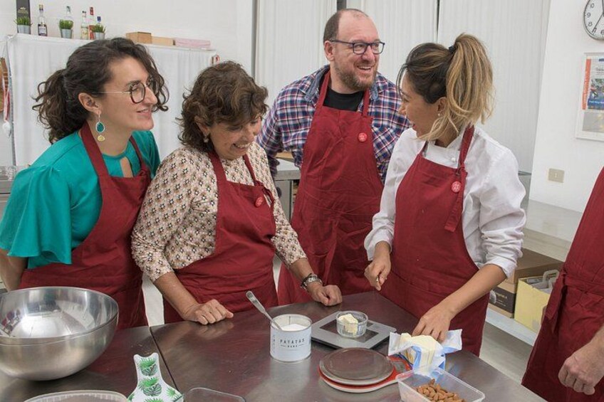 Een groep mensen met schorten, bezig met koken, met kommen, maatbekers en ingrediënten op een tafel. 1