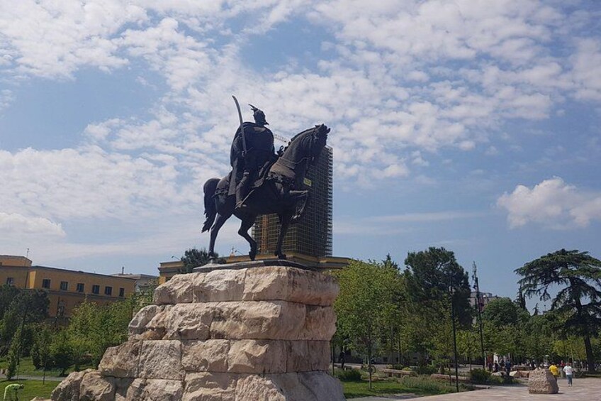 Une statue représentant une personne à cheval, placée sur un piédestal dans un parc, avec des arbres et des bâtiments en arrière-plan. 3
