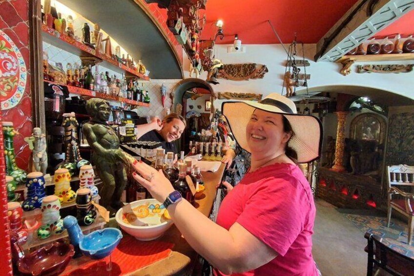 Una persona con camisa rosa y sombrero de ala ancha en un bar, con varias botellas y una estatua verde. 3