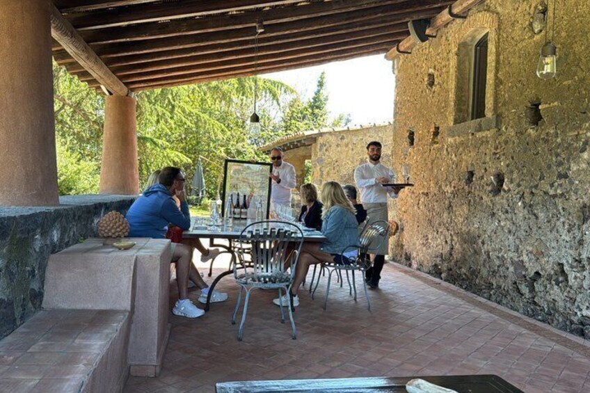 En grupp människor sitter och äter vid ett utomhusbord, och en servitör står i närheten. I bakgrunden syns en rustik stenmur och en träpergola. 4