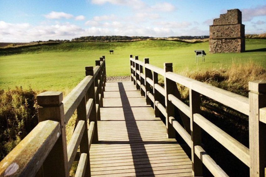Un pont en bois menant à un terrain de golf, avec une structure en pierre visible au loin. 2