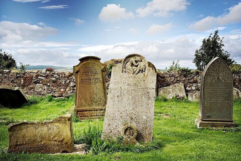 A cemetery with stone headstones, some with inscriptions, set against a backdrop of a stone wall and grassy area. 3