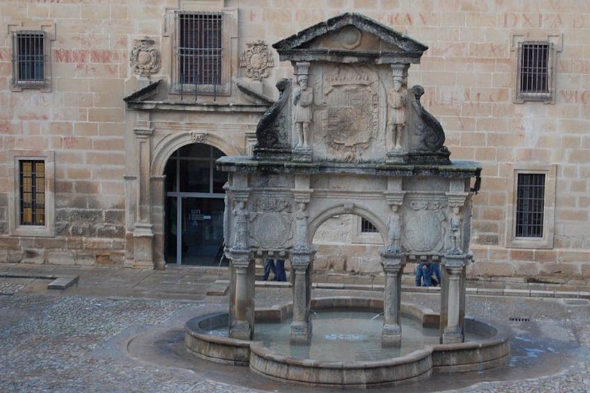 A historical fountain with intricate carvings, a stone building with arched windows, and a cobblestone ground. 1