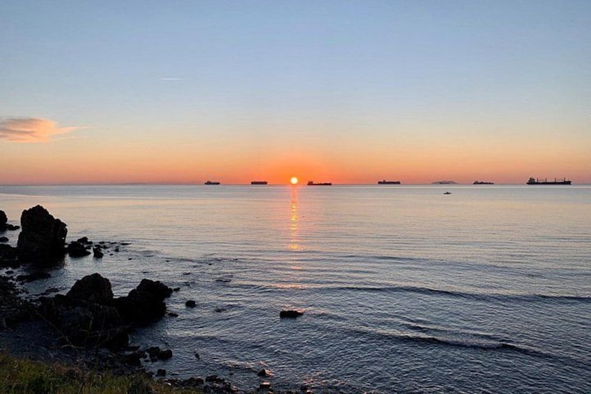 A sunset over a calm sea with rocks on the shore and distant ships. 2