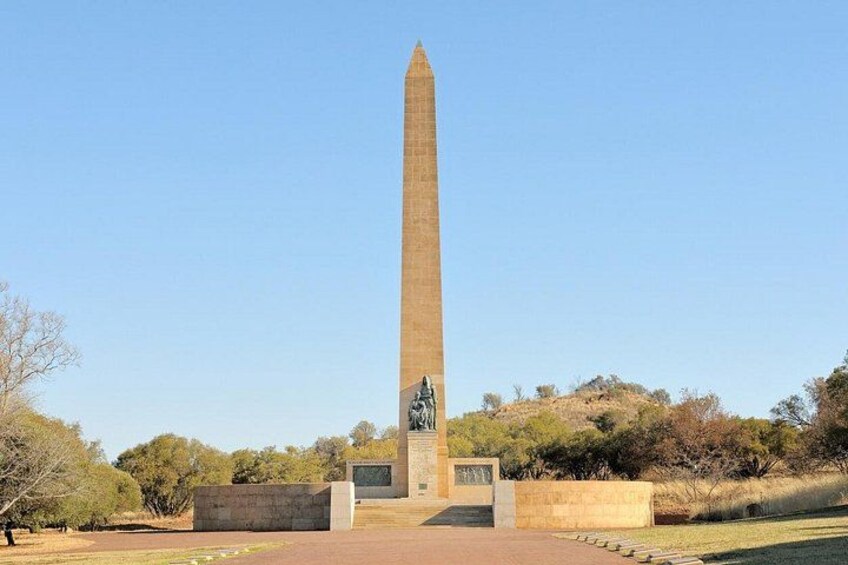 A tall obelisk monument with a pedestal base, surrounded by a landscaped park with sparse trees and a clear sky. 5