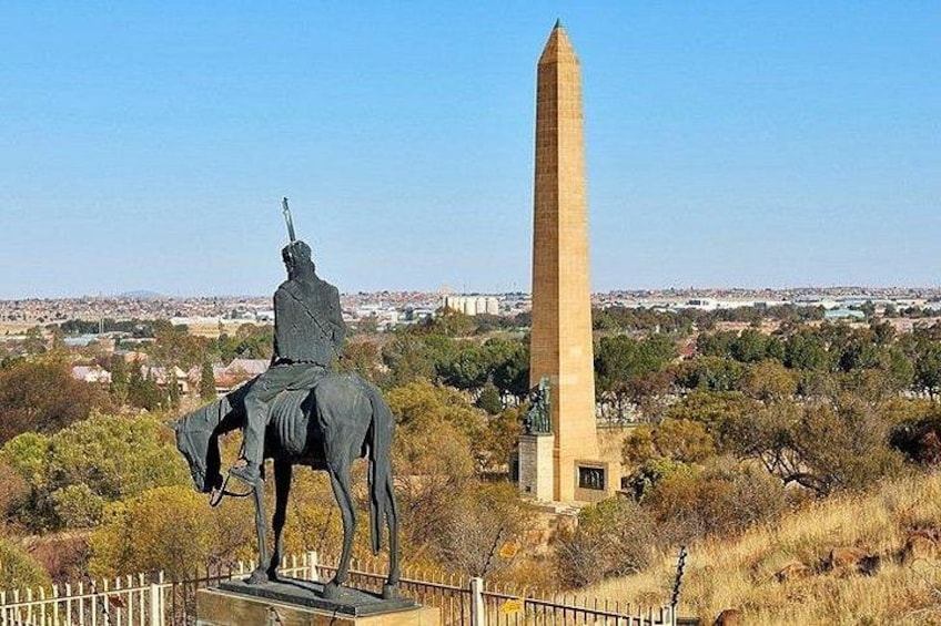 Een standbeeld van een zittende figuur op een paard, een hoge obelisk, en een omheind gebied met bomen op de achtergrond. 1