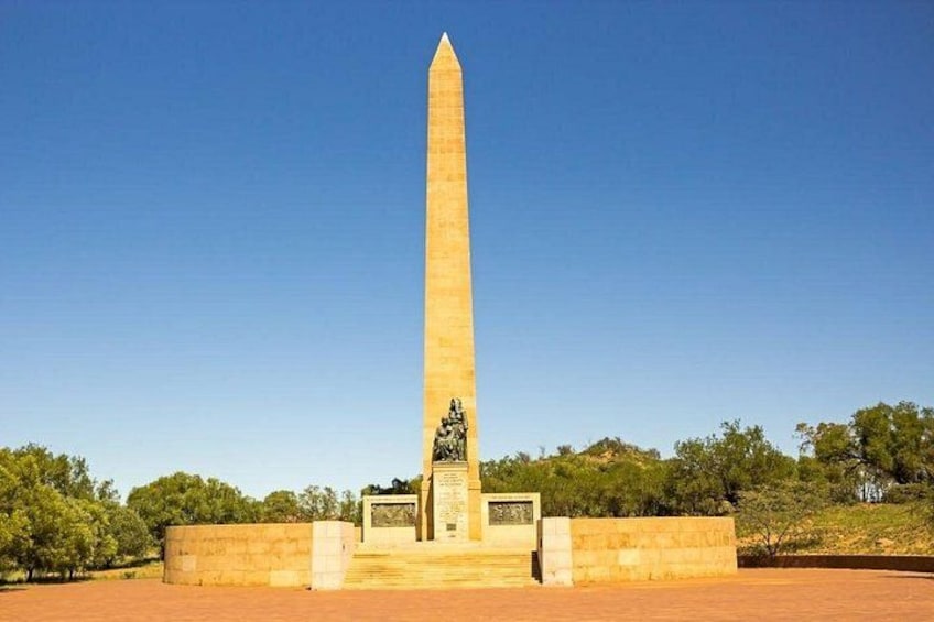 Een hoge, slanke obelisk met een voetstuk, omgeven door een lage muur en groen. 4