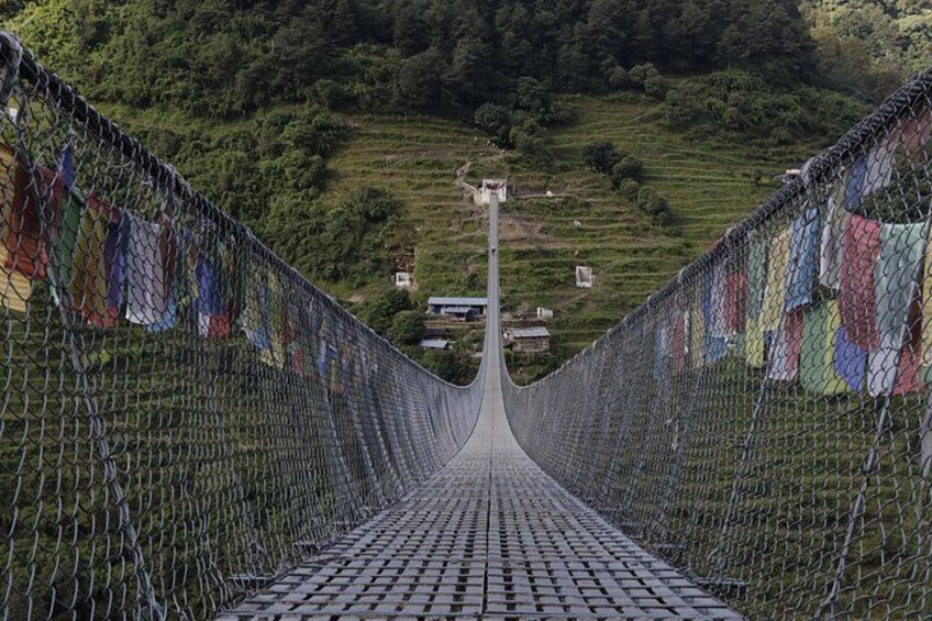 A suspension bridge with colorful prayer flags on both sides, leading to a village in a hilly landscape. 2