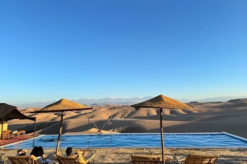 A desert landscape with a swimming pool, lounge chairs, and thatched umbrellas. 5