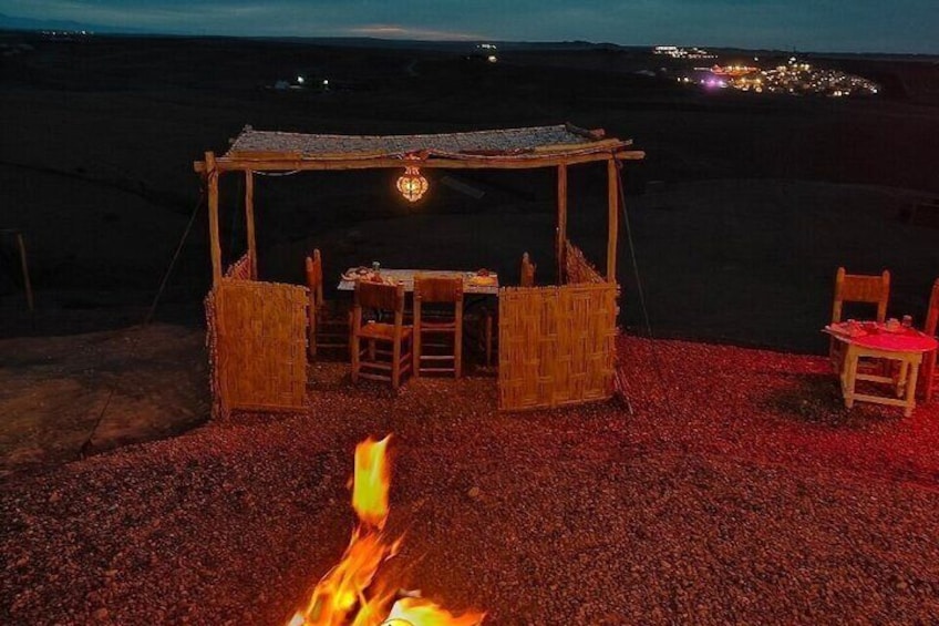 A rustic outdoor dining setup with a fire in the foreground, wooden chairs, and a table under a thatched roof. 5