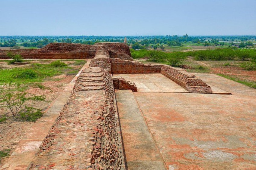 Ancient ruins with stepped structures and a flat area, surrounded by open fields and distant trees. 5