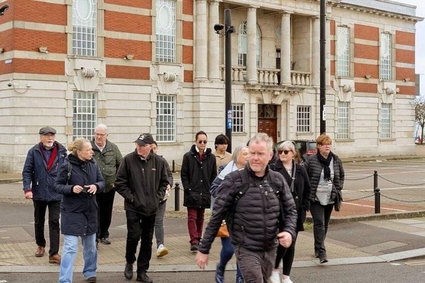 A group of people walking on a sidewalk in front of a large building with columns and a brick facade. 3