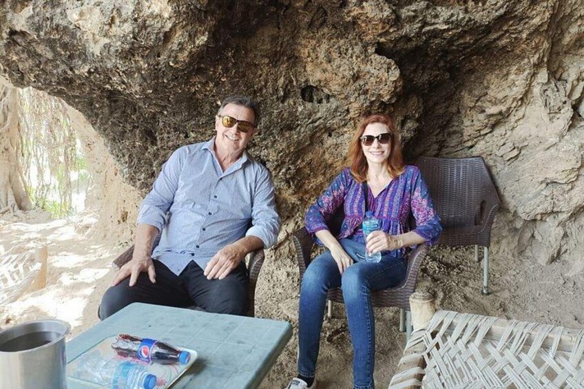 Two people sitting on wicker chairs under a rocky overhang, with a table and a metal container in front of them. 2