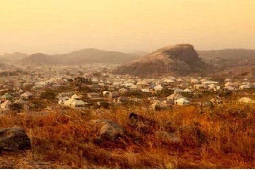 A landscape with a prominent rock formation, a town in the background, and a field of dry grass in the foreground. 2