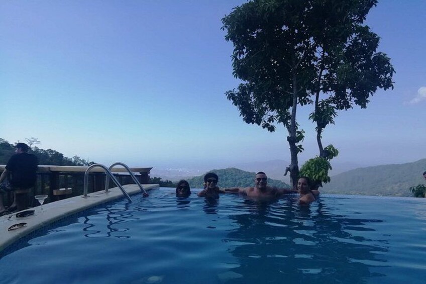 A swimming pool with people relaxing, surrounded by trees and a mountainous landscape. 2