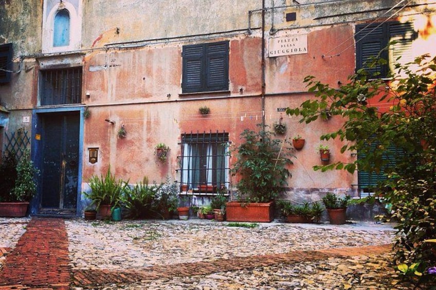 A cobblestone courtyard with potted plants, a brick pathway, and a building with a sign reading 'PIAZZA DEI BIANCHI.' 3