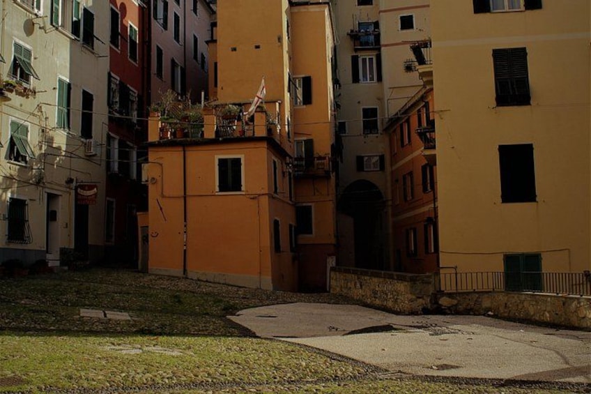 A narrow street with buildings on either side, a small grassy area, and a paved path. 2