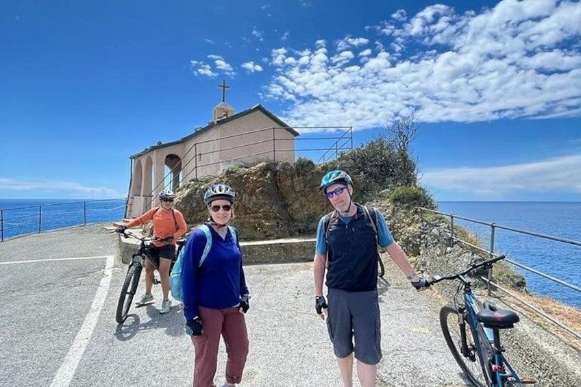Trois cyclistes sur une route côtière, avec une église et l’océan en arrière-plan. 2