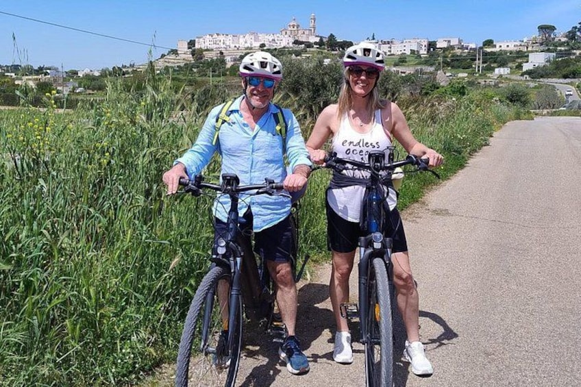 Due persone in bicicletta su una strada, con vegetazione e edifici sullo sfondo. 2
