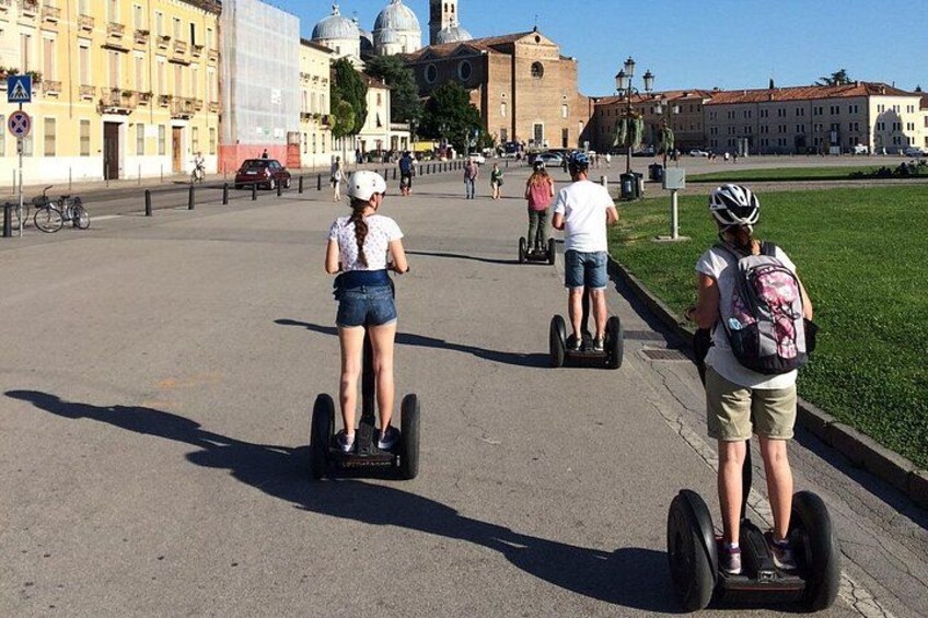 Personas montando Segways por un camino pavimentado en una zona urbana, con edificios y un campo de césped al fondo. 3