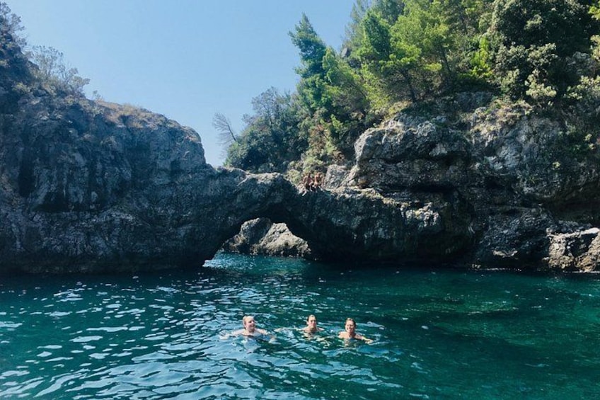 A natural rock formation with a water arch, clear blue water, and surrounding greenery. 5