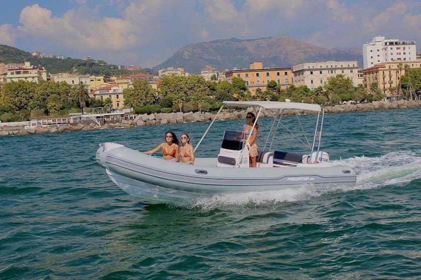 A motorboat on water with two people on board, a coastal town in the background, and mountains in the distance. 1