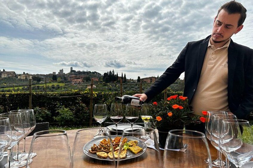 A man serving food at an outdoor table with a vineyard and town in the background. 1