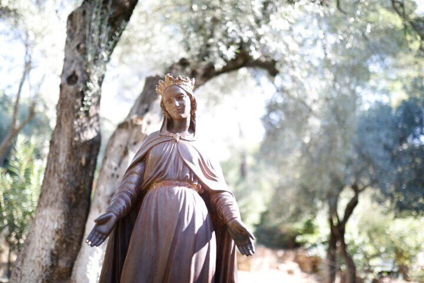 A statue of a robed figure with a crown, standing in a wooded area with trees and sunlight filtering through. 4