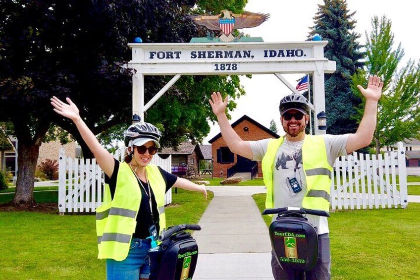 Two people with raised arms in front of a sign that reads "FORT SHERMAN, IDAHO. 1878". 2